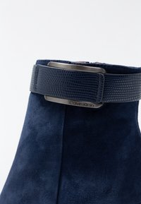 Marineblauwe suède enkellaars met een gestructureerde marineblauwe riem en een rechthoekige metalen gesp met "Calvin Klein"-branding.