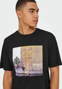 Camiseta de algodón negra con un estampado gráfico a color de una flor y una figura sentada en una calle, enmarcada por un diseño rectangular en contraste.