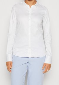 Camicia bianca a maniche lunghe con colletto classico, caratterizzata da una texture liscia e un orlo curvo per una vestibilità aderente.