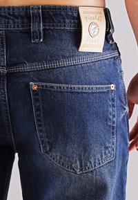 Dunkelblaue Denim-Jeans mit strukturierter Oberfläche, ausgestattet mit einem Lederpatch an der Taille, Doppelnaht und einer Gesäßtasche mit Kupfernieten-Akzenten.