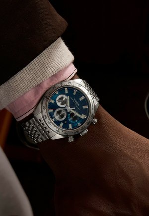 Montre chronographe en acier inoxydable avec un cadran bleu, des accents argentés et trois sous-cadrans ; portée sur un poignet présentant une surface texturée marron.