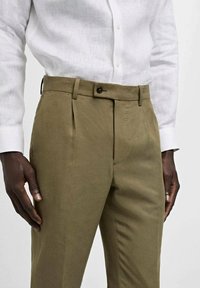 Olive green trousers with a smooth texture, featuring a button closure and pleats. Paired with a white button-up shirt.