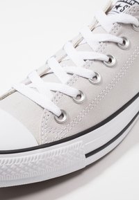 Baskets en toile gris clair avec lacets blancs, œillets argentés et embout en caoutchouc. Elles présentent une semelle extérieure blanche texturée avec des accents noirs.