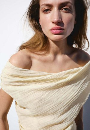 Jeune femme à la peau claire et aux cheveux bruns détachés, portant un haut crème texturé à épaules dénudées sur fond blanc.
