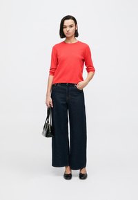 Parte superior roja de algodón de manga larga combinada con jeans de mezclilla oscuros de pierna ancha. El conjunto se completa con flats de ballet negros y un bolso pequeño negro.