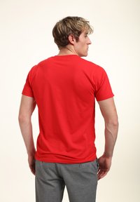 Červené tričko s krátkým rukávem a kulatým výstřihem, vyrobené z bavlny. Výrazně hladká textura, přiléhavý design. Nosené s šedými kalhotami, zobrazené zezadu.