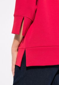 Felpa rossa con maniche a tre quarti, caratterizzata da un dettaglio con fessura laterale e una vestibilità rilassata, abbinata a jeans blu scuro.