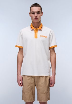 Valkoinen polo-paita oranssilla kauluksella ja hihoilla, rintataskussa logo, yhdistettynä vaaleanruskeisiin shortseihin. Silkkinen puuvillapinta hienovaraisilla yksityiskohdilla.