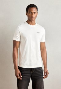 Witte katoenen T-shirt met een ronde hals en korte mouwen. Bevat een klein bedrukt logo op de linkerborst. Gecombineerd met een donkere spijkerbroek.