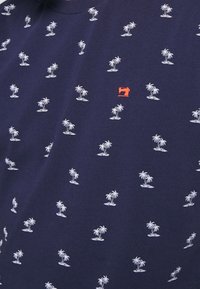 T-shirt blu navy con un motivo ripetuto di piccole palme bianche e un logo di macchina da cucire rossa sul petto. Materiale in cotone.