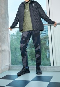 Svart quiltad jacka, olivgrön isolerad tröja, joggers med camouflage-mönster i marinblått och grått, samt svarta sportskor. Modern urban outfit.