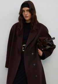 Bruine wollen jas met een losse pasvorm, voorzien van grote knopen, een brede kraag en een gestructureerde stof. Draagt over een zwart outfit en houdt een tas vast.