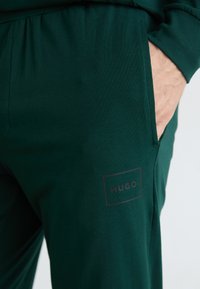 Person som har på seg mørkegrønne joggebukser med "HUGO"-logo på låret, med hånden avslappet plassert i sidelommen.