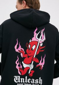 Person iført sort hættetrøje med rød tegneserie-djævel med pink flammer, der holder en trefork, og teksten "Slip din indre djævel løs."