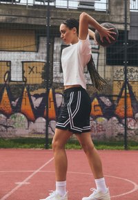 Black shorts with white side stripes, paired with a white short-sleeve shirt and white sneakers. Basketball held overhead. Urban court backdrop.