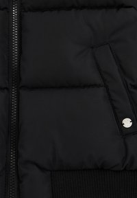 Doudoune noire avec fermeture éclair, texture matelassée et ourlet en tricot côtelé. Comprend un détail en bouton argenté sur la poche avant.