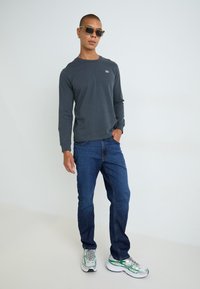 Langermet mørkegrå skjorte med en liten logo, kombinert med blå jeans og grå og grønne joggesko; solbriller fullfører antrekket.