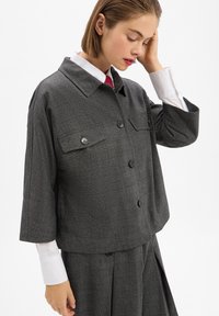 Grau kariertes Cropped-Jackett mit Spitzkragen, zwei Brusttaschen und Knopfverschluss. Über einem weißen Hemd mit roter Krawatte getragen.