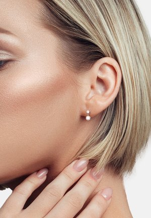 Silberne Tropfenohrringe mit einem kleinen runden Stecker und einer größeren Perle, die darunter baumelt, vor glatter Haut und kurzen blonden Haaren.