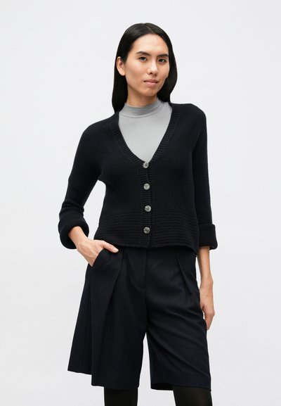 Tengerészkék kötött kardigán V-nyakkivágással, öt gombos záródással és felhajtott ujjakkal, elegáns tengerészkék rövidnadrággal párosítva. Sima textúra, laza szabás.