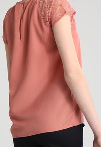 Bladoróżowa bluzka z krótkimi koronkowymi rękawami i otworem w tylnej części. Gładka tekstura, luźny krój oraz zaokrąglony dół.