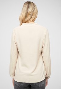 Maglia lungo maniche beige chiaro con scollatura rotonda e una finitura liscia, leggermente texture, abbinata a jeans grigi. Nessun motivo visibile.