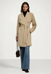 Žena v béžovom kabáte s opaskom, čiernom roláku a širokých modrých džínsach, so malou hnedou kabelkou s reťazovou rúčkou, stojí na bielom pozadí.