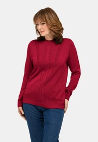 Frau mit schulterlangem braunem Haar, die einen roten Strickpullover mit gemustertem Vorderteil und blaue Jeans trägt, und nach links unten schaut.