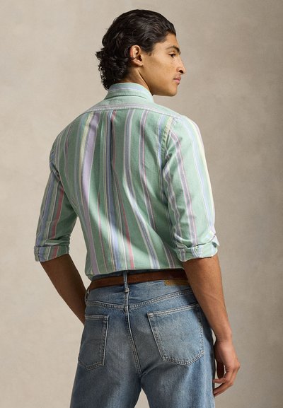 Muškarac okrenut bočno, nosi prugastu pastelnu košulju s podvrnutim rukavima uvučenu u plave traperice sa smeđim remenom na neutralnoj pozadini.