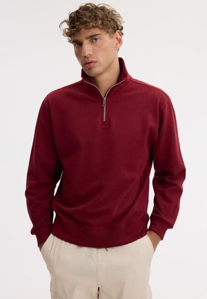 Un joven posa con una sudadera burdeos con cremallera corta y unos pantalones beige sobre un fondo liso y claro.