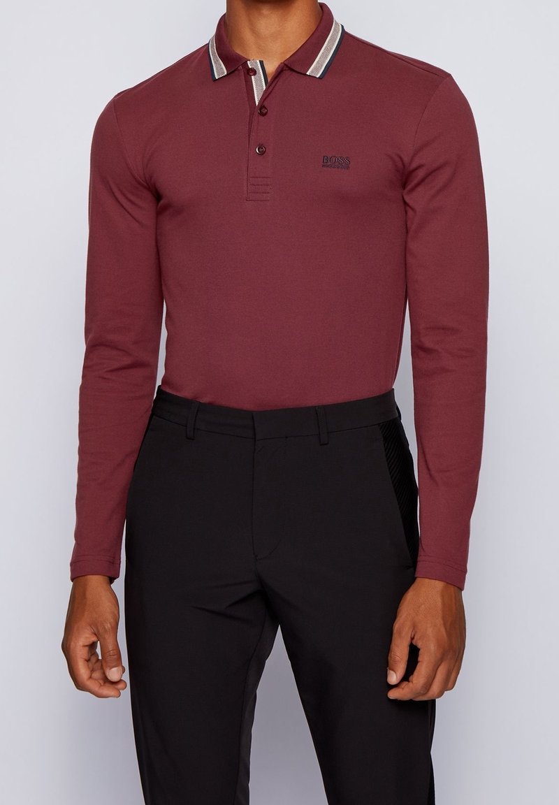 Burgundy långärmad polo med randig krage, med en logotyp på bröstet och i kombination med svarta skräddarsydda byxor.