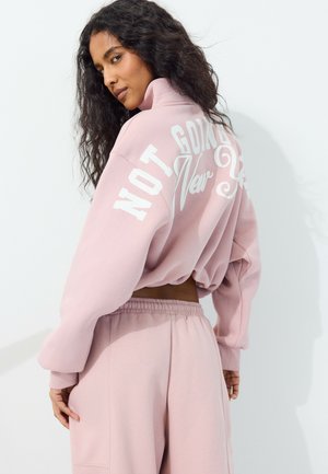 Kvinde med langt krøllet hår iført en lyserød cropped sweatshirt og bukser, kigger over skulderen, tekst på sweatshirten læses "Not Going" synlig.