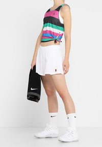 Kleurvolle gestreepte tanktop in roze, groen en zwart, gecombineerd met witte shorts. Model houdt een zwarte handdoek vast en draagt witte sokken en schoenen.