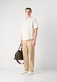  Lichtbeige kortekrans overhemd met verticale strepen, gecombineerd met kaki broeken en witte sneakers. Houdt een donkergroene tas vast.