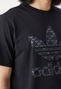 Černé bavlněné tričko s velkým grafickým logem zobrazujícím vzorovaný list, zvýrazněné horizontálními pruhy v tmavším odstínu.