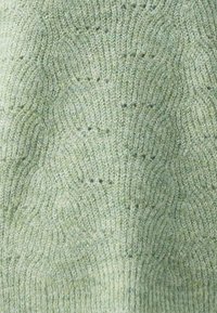 Strukturierter Strickstoff in sanften Grüntönen, mit einem Wellenmuster und verschiedenen Schattierungen, der eine gemütliche, warme Ausstrahlung hat.