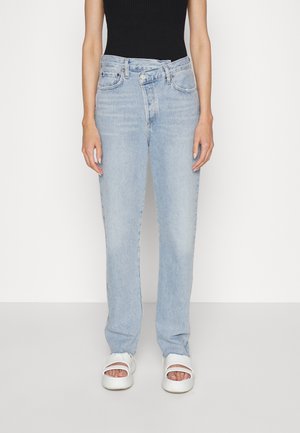 Person trägt hellblaue High-Waist-Jeans mit überlappendem Knopfdetail, schwarzes ärmelloses Top und weiße Plateausandalen mit offenem Zehenbereich, steht.