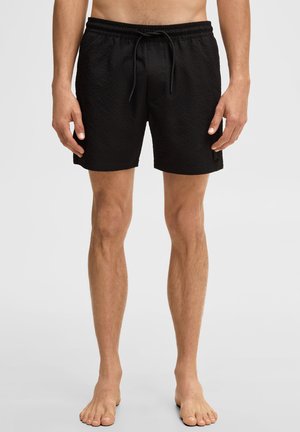 Man draagt zwarte gestructureerde zwemshorts met elastische tailleband en trekkoord, staat blootsvoets tegen een effen achtergrond.