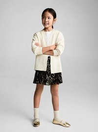 Witte gebreide cardigan over een wit shirt, gecombineerd met een zwarte geplooide rok met gouden stippen. Draagt beige sokken en gouden ballerina's.