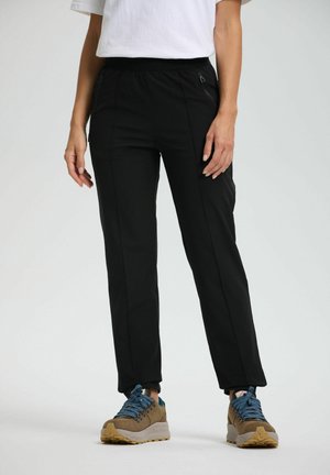 Persoon in zwarte taps toelopende broek met ritszakken en bruine wandelschoenen met blauwe veters, staande tegen een effen lichte achtergrond.