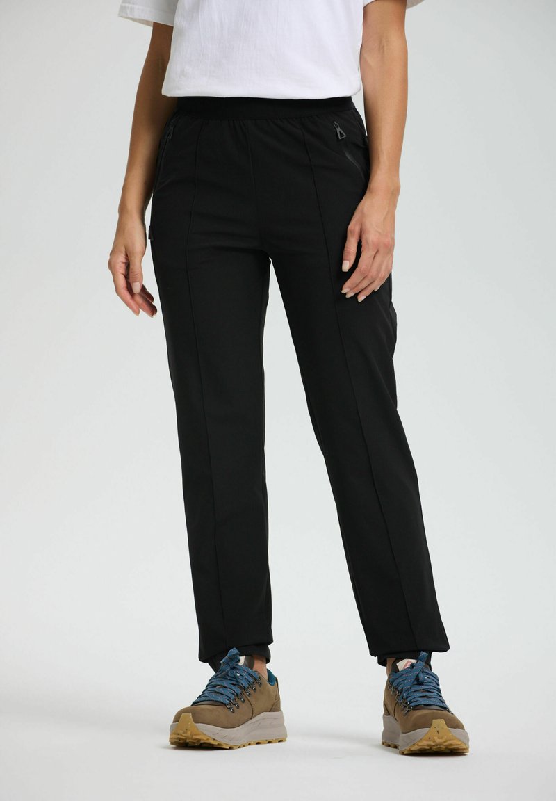 Henkilö, jolla on päällä mustat kapeat housut vetoketjupusseilla ja ruskeat vaelluskenkä sinisillä nauhoilla, seisoo yksinkertaista vaaleataustaa vasten.