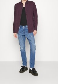 Rutig maroon skjorta över en svart linne, bärs med slitna blå jeans och svarta skor med fransdetalj.