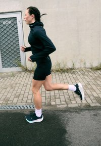 Camisola de corrida de manga longa preta, calções pretos, meias brancas até ao tornozelo e sapatilhas de corrida pretas com detalhes verdes; a correr num caminho pavimentado ao ar livre.