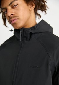 Jachetă neagră cu fermoar și glugă, având o textură netedă și logo-ul "Dickies" pe piept, evidențiind un design minimalist și accesorii funcționale.