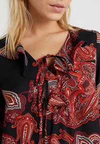 Svart blus med intrikat röd paisleymönster, krage med volanger och snörning framtill. Slät textur och lättviktsmaterial.