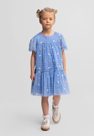 Jeune fille aux cheveux blonds coiffés en deux chignons, portant une robe bleue à fleurs et à pois, des chaussettes blanches à volants et des baskets beige, debout sur un fond uni.