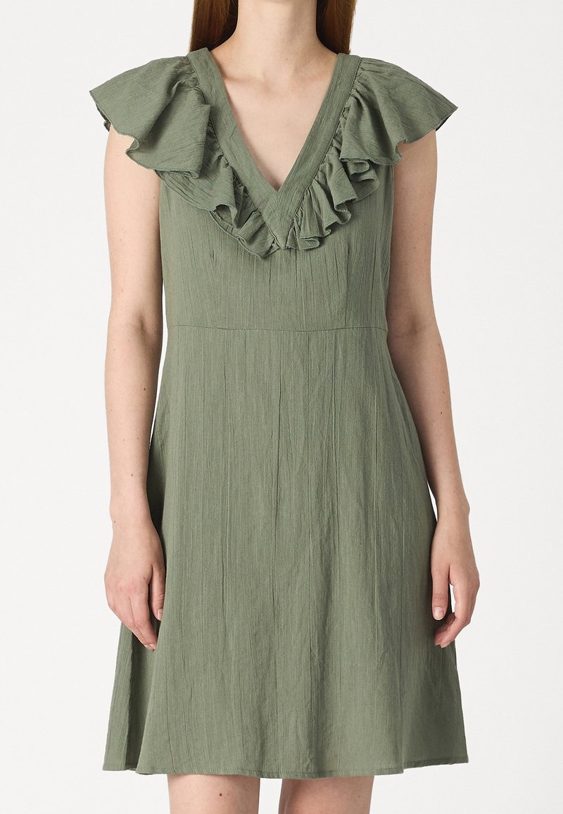 Robe sans manches vert olive avec un décolleté en V et un volant autour du cou et des épaules, portée par une personne à la peau claire.