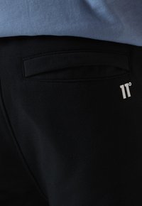 Los pantalones de chándal negros tienen un bolsillo trasero y un pequeño logo blanco. El material parece suave y de textura lisa.