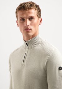 Jonge man met kort krullend haar, in een lichtgrijze gebreide trui met een halve rits kraag en een zwarte hexagonale patch op de mouw.
