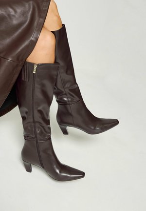 Femme portant des bottes en cuir marron foncé montant jusqu'aux genoux avec des talons bas et une jupe en cuir assortie sur un fond clair uni.